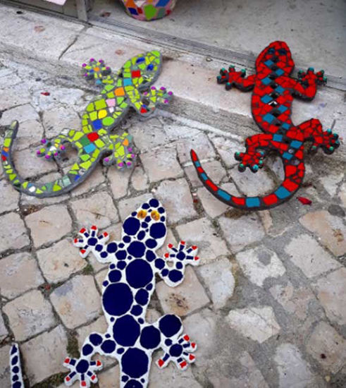 Three colorful mosaic geckos made of ceramic tiles, each with unique designs, arranged on the ground in a vibrant display.